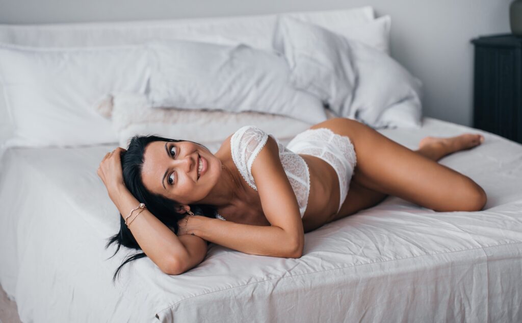 young tanned woman in white lingerie laying on bed at home toothy smiles, satisfied by body shape